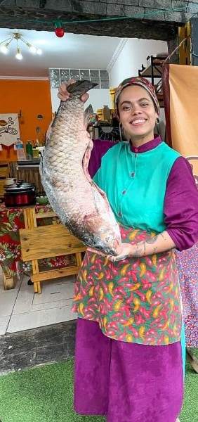 Chef Bia Souza sorrindo, de lenço e avental colorido, segurando um peixe grande