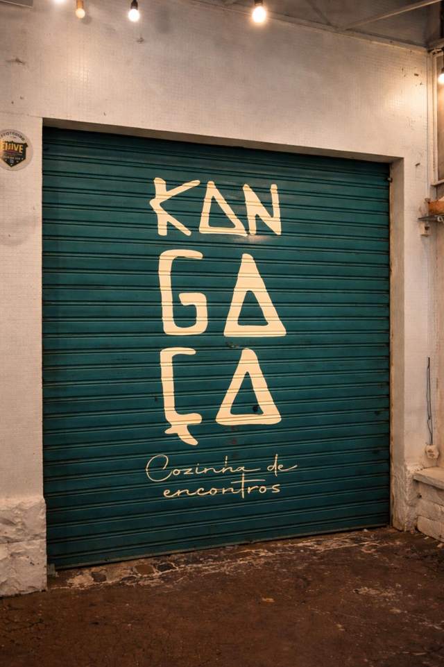 Porta de aço turquesa do Kangaça, com o logo e a assinatura Cozinha de encontros pintados em bege, iluminada à noite